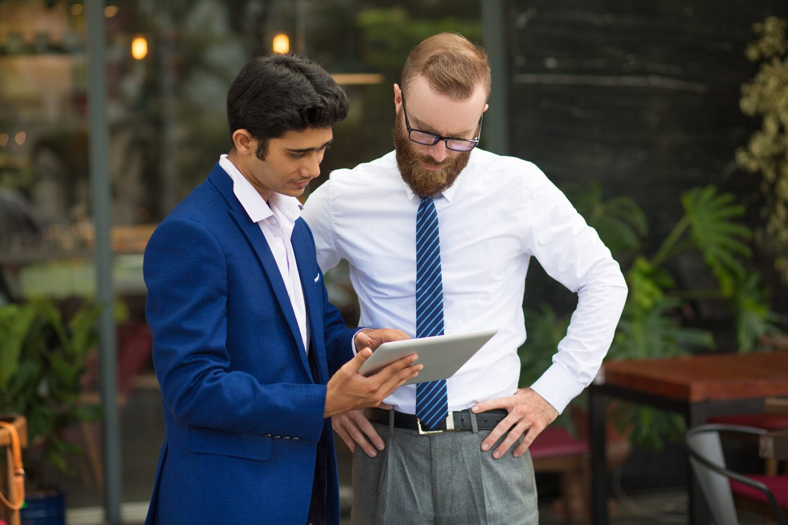 Pensive concentrated managers standing in modern cafe and analyzing online data. Indian assistant in formalwear showing report to bearded executive. Technology concept