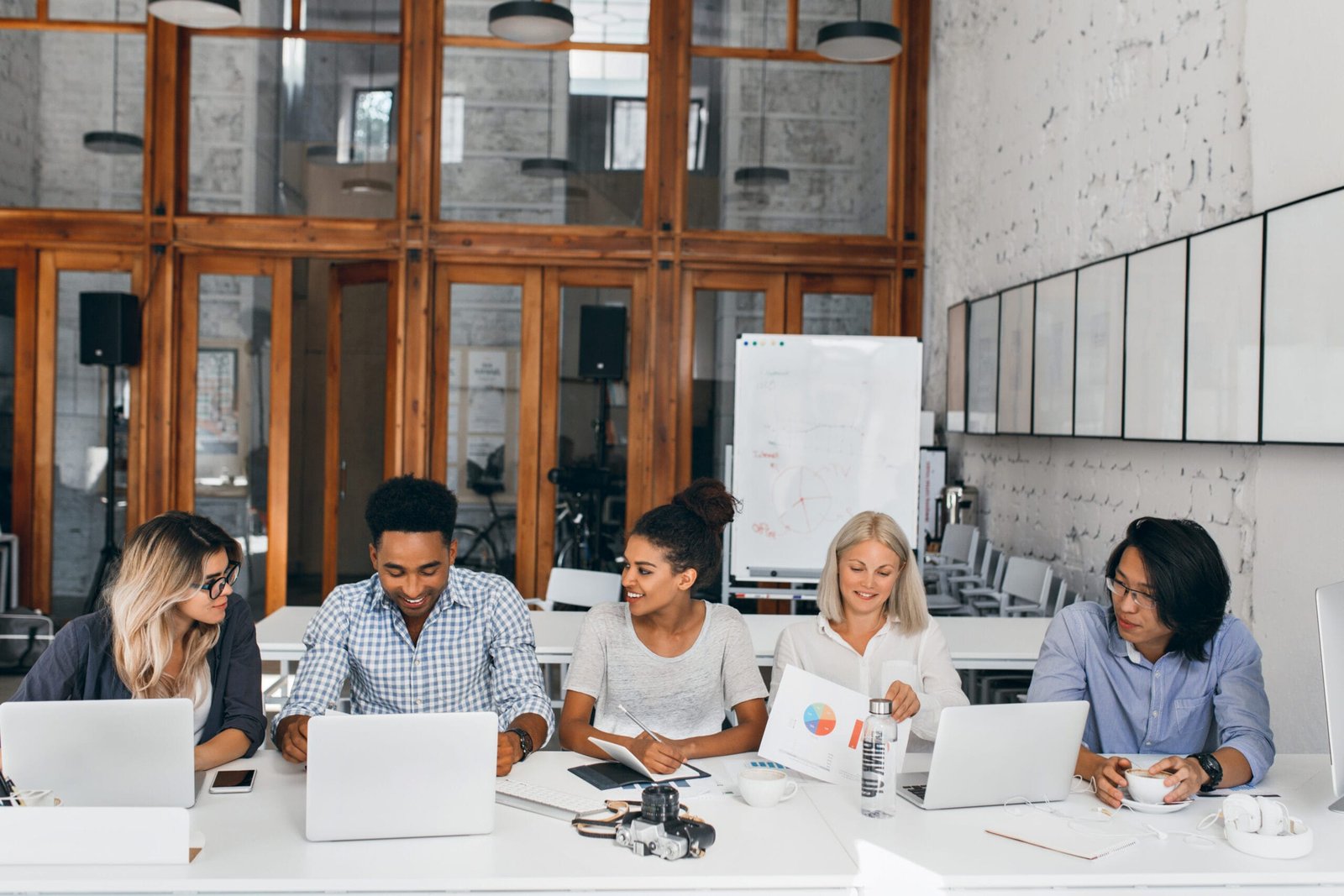 Tired asian it-specialist drinking coffee and watching female colleague working with laptop. Indoor portrait of young business people sitting at the table together in conference hall.