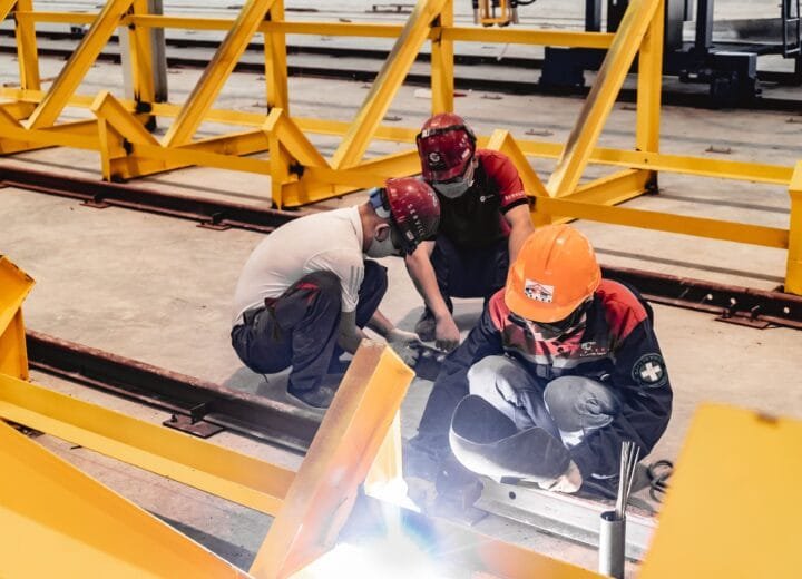 Three construction workers welding metal structures indoors, wearing safety gear