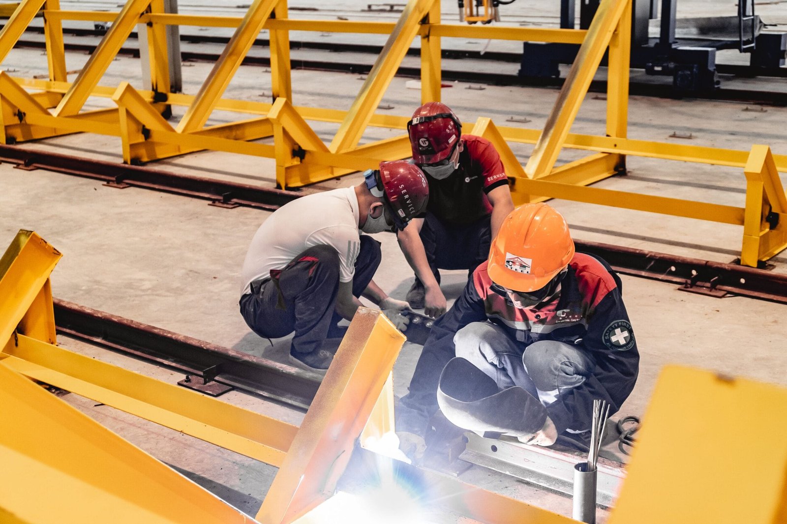 Three construction workers welding metal structures indoors, wearing safety gear