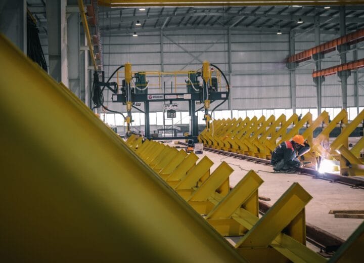 A worker welding metal in an industrial warehouse with heavy machinery