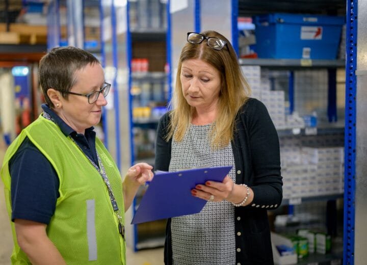 Two women in a warehouse reviewing an inventory checklist on a clipboard